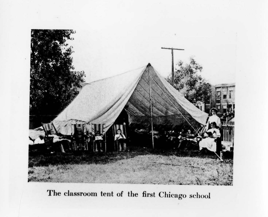 black and white photograph of an outdoor tent. children sit in folding chairs outside it, alongside their teacher, who holds up an open book.
